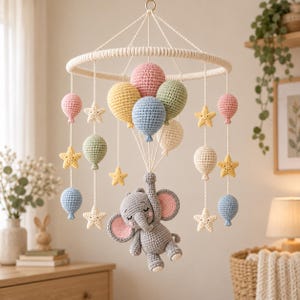 Puede incluir: Un móvil de ganchillo con un elefante gris central con orejas rosas, suspendido por hilos blancos de un marco circular. El móvil incluye varios globos de ganchillo en tonos pastel rosa, amarillo, azul y verde, junto con pequeñas estrellas amarillas y blancas.