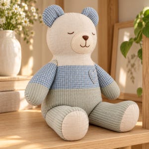 Pode incluir: Um urso de peluche com cabeça cor de creme, olhos fechados bordados e nariz castanho. O tronco apresenta um padrão axadrezado azul e branco com um pequeno coração, enquanto os braços e as pernas têm riscas verdes e brancas.