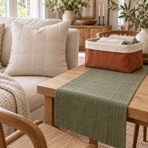 Peut inclure: Un panier de rangement rectangulaire en tissu avec une base couleur rouille et un bord supérieur crème, contenant des textiles pliés beiges et verts. Le panier est posé sur une table en bois recouverte d'un chemin de table vert sauge.