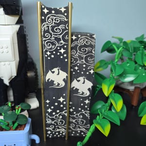 Op de afbeelding: Twee boeken met zwarte paginaranden voorzien van een wit zeefdrukontwerp van draken, wolken en sterren. De boeken hebben goudkleurige ruggen en staan verticaal opgesteld, waardoor de decoratieve kunst op de snede duidelijk zichtbaar is.