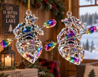 Pode incluir: Dois ornamentos de cristal facetado iridescente em forma de pessoas em caiaques, cada uma segurando um remo com pás de cores arco-íris. O caiaque à esquerda apresenta o nome Noah e o caiaque à direita apresenta o nome Chloe.