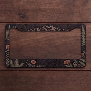 May include: A rectangular black license plate frame featuring an engraved design of mountain peaks along the top edge, tall pine trees on the sides, and various wildflowers and fern leaves along the bottom and side borders with topographic lines.