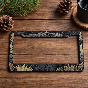May include: A rectangular black number plate frame featuring gold-toned line art illustrations. The design includes mountain ranges, evergreen trees, ferns, wildflowers, and topographical map lines across the top and bottom borders.