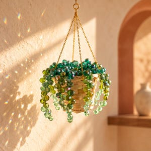 May include: A hanging plant decoration featuring strands of faceted green and teal glass beads arranged to resemble a string of pearls. The beads are contained within a small, light-colored woven basket suspended by a thin gold-toned metal chain.