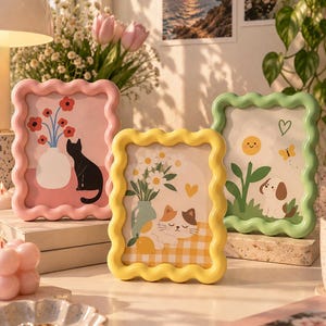 Puede incluir: Tres marcos de fotos rectangulares con bordes ondulados en rosa pastel, amarillo y verde claro. El marco rosa muestra un gato negro junto a un jarrón con flores rojas. El marco amarillo presenta un gato calicó descansando junto a margaritas. El marco verde incluye un cachorro, una flor sonriente y una mariposa.