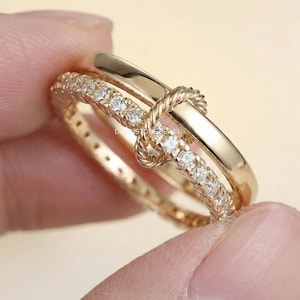 Könnte beinhalten: Ein goldener Ring mit einem Doppelband-Design, bei dem ein Band glatt und poliert ist, während das andere mit einer Reihe kleiner, klarer, rund geschliffener Steine besetzt ist. Die beiden Bänder sind durch einen zentralen, gedrehten Goldknoten verbunden.