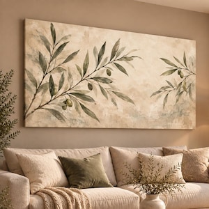 Op de afbeelding: Een groot rechthoekig wandkunstwerk met een aquarelachtige schildering van olijftakken met groene bladeren en kleine olijven tegen een getextureerde beige achtergrond, opgehangen boven een beige bank met diverse kussens en een plaid.