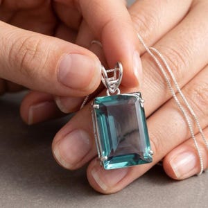 May include: A rectangular, faceted teal gemstone pendant set in a silver-toned metal prong setting. The pendant hangs from a delicate silver-toned chain. The gemstone displays a deep, translucent blue-green hue with sharp, clean edges.