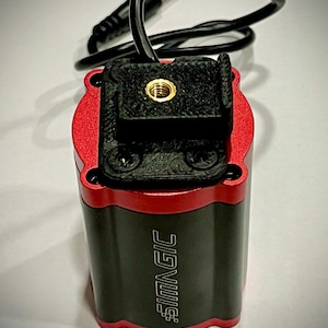 Puede incluir: Un dispositivo electrónico cilíndrico de color negro y rojo con el texto SIMAGIC impreso verticalmente en el lateral. La parte superior cuenta con un soporte negro impreso en 3D con una inserción roscada de latón centrada y un cable negro.