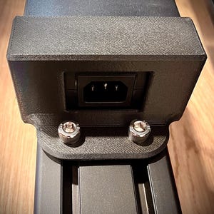 Puede incluir: Un soporte de fuente de alimentación negro impreso en 3D con una carcasa rectangular y un puerto de entrada de alimentación C14 centrado. El soporte está fijado a un riel de extrusión de aluminio negro con dos pernos plateados hexagonales.