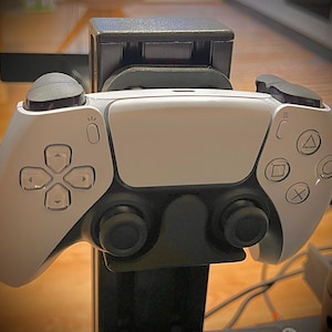 Op de afbeelding: Een wit en zwart draadloze videogamecontroller gemonteerd op een zwarte verticale standaard. De controller is voorzien van een centraal touchpad, twee analoge sticks, een richtingspad en vier actieknoppen met driehoek-, cirkel-, kruis- en vierkantsymbolen.
