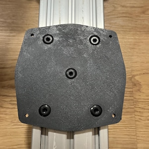 Peut inclure: Une plaque de montage gris foncé, texturée et en forme de bouclier, fixée sur un rail d'extrusion en aluminium argenté. La plaque comporte cinq boulons hexagonaux noirs disposés en motif, avec deux trous circulaires vides aux coins.
