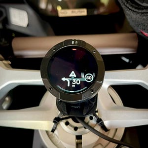 Op de afbeelding: Een rond zwart digitaal navigatiescherm gemonteerd op een zilverkleurige motorstuurconstructie. Het scherm toont een witte afbeelding met een pijl naar links, de tekst 30 m en een cirkelvormig icoon met het getal 50 op een donkere achtergrond.