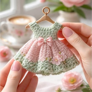 Pode incluir: Um vestido de crochê em miniatura em verde pastel e rosa, pendurado num pequeno cabide de madeira. O vestido apresenta um laço de cetim rosa na cintura e dois pequenos adornos florais rosa com centros de pérolas na saia. O texto diz ViTaSjaCraft craft bases & supplies.