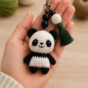Può includere: Un portachiavi a forma di panda all'uncinetto con un piccolo orso panda in filato bianco e nero, perline di plastica nera, una sfera lavorata all'uncinetto bianca e una nappa in tessuto verde scuro fissata a un moschettone in metallo bronzo.