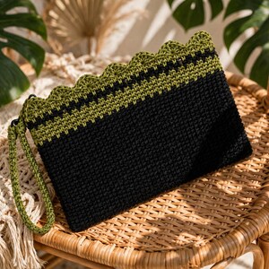 Puede incluir: Un bolso de mano rectangular de ganchillo con una sección principal negra, una banda horizontal de color verde oliva y un borde superior festoneado en verde oliva. El bolso incluye una correa de muñeca a juego en verde oliva.