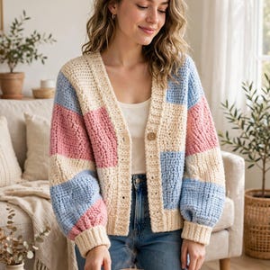 May include: A chunky crochet cardigan featuring a colour-blocked pattern of cream, light blue, and soft pink squares. The garment has a V-neckline, long sleeves with ribbed cuffs, and a single brown button closure at the centre front.