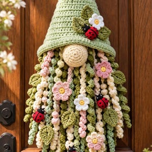 Peut inclure: Une décoration murale gnome en crochet avec un grand chapeau vert sauge, un nez beige rond et une longue barbe faite de brins de laine en spirale dans des tons de vert, crème et rose. Le design inclut des fleurs et des coccinelles rouges.