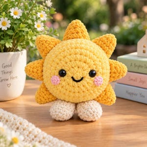 Op de afbeelding: Een geel gehaakt zon-knuffeltje met een rond gezicht, zwarte kraaloogjes, een geborduurde zwarte glimlach en roze wangen. De zon heeft gestructureerde stralen rondom de omtrek en twee kleine, ronde, crèmekleurige voetjes aan de onderkant.