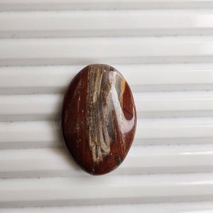 May include: An oval-shaped polished gemstone cabochon featuring a deep reddish-brown base color with prominent vertical streaks of cream, tan, and dark brown mineral inclusions running through the center, creating a natural wood-like appearance.