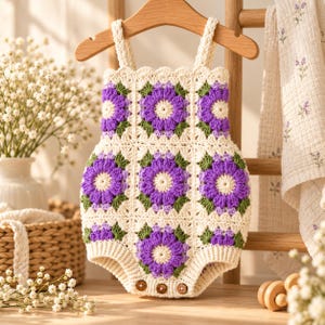 May include: A sleeveless crochet romper featuring a granny square pattern with cream-colored borders and central purple flower motifs with green leaves. The garment has a scalloped neckline, two shoulder straps, and three small brown buttons at the base.