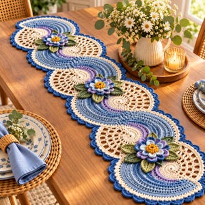 Può includere: Un lungo runner da tavola all'uncinetto con un motivo a onde ripetuto nei toni del blu, viola e crema. Il runner è decorato con fiori tridimensionali a più strati nei colori blu e giallo, con foglie verdi applicate a intervalli regolari.