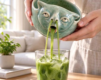 May include: A light green ceramic bowl shaped like a cat face with a wide mouth and nostrils. The bowl is tilted to pour a vibrant green liquid into a clear glass filled with ice cubes, creating a steady stream from the cat's nose and mouth.
