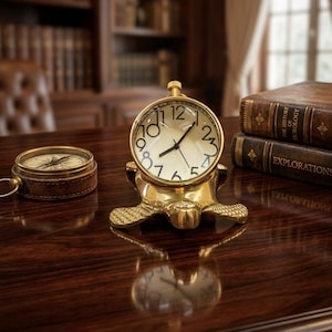 Könnte beinhalten: Eine schreibtischuhr aus messing in form einer schildkröte mit rundem zifferblatt, schwarzen zeigern und schwarzen ziffern. Neben der uhr liegt ein runder messingkompass mit braunem ledergehäuse. Die uhr hat einen aufziehknopf an der oberseite.