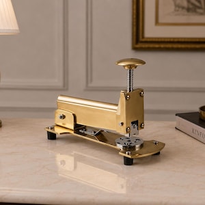 May include: A gold-toned metal stapler featuring a vertical plunger mechanism with a circular top handle, a rectangular body, and a wide base supported by four small black rubber feet. The device includes a circular metal plate at the front.