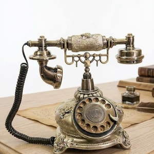 Könnte beinhalten: Ein antikes Drehscheibentelefon mit metallisch bronzefarbener Oberfläche, das auf der Basis und dem Hörer mit detaillierten Blumenreliefs verziert ist. Das Gerät verfügt über eine kreisförmige Wählscheibe mit Zahlen und ein schwarzes Spiralkabel.