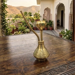 May include: A polished brass hookah base featuring an ornate floral embossed pattern on the lower bulbous section, a vertical stem with a wide bowl at the top, and a single angled tube extending from the side, all crafted from shiny metallic brass.