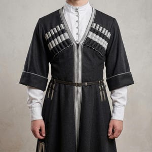 May include: A dark gray long-sleeved coat featuring a white high-neck shirt underneath. The coat has silver trim, decorative cartridge loops across the chest, and a thin dark belt with silver accents at the waist, creating a structured appearance.