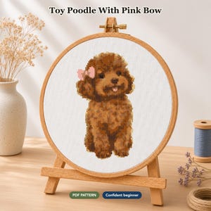 Op de afbeelding: Een kruissteekproject in een ronde houten borduurring op een kleine houten ezel. De stof toont een bruine toy poedel met een roze strik. De tekst bovenaan luidt Toy Poodle With Pink Bow, met de labels PDF PATTERN en Confident beginner.