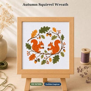 Op de afbeelding: Een kruissteekpatroon getiteld Autumn Squirrel Wreath met twee oranje eekhoorns omringd door een cirkelvormige krans van groene en oranje eikenbladeren en eikels, gepresenteerd in een lichthouten lijst op een houten ezel.