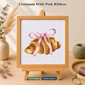 Op de afbeelding: Een kruissteekpatroon met een goudbruine croissant gestrikt met een lichtroze lint, weergegeven in een vierkante houten lijst op een kleine ezel. De tekst Croissant With Pink Ribbon, PDF PATTERN en Confident beginner is zichtbaar.