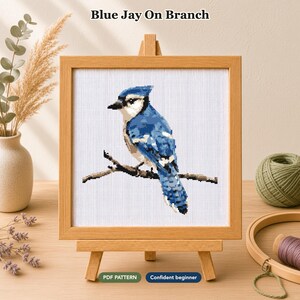 Op de afbeelding: Een kruissteekpatroon van een blauwe gaai op een tak, gepresenteerd in een lichthouten lijst op een kleine ezel. De tekst "Blue Jay On Branch" staat erboven, met de labels "PDF PATTERN" en "Confident beginner" aan de onderzijde.