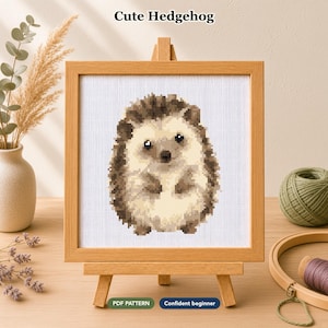Op de afbeelding: Een kruissteekpatroon met een gepixelde egel in bruin- en crèmetinten op witte stof, gepresenteerd in een lichthouten lijst op een kleine ezel. De tekst "Cute Hedgehog", "PDF PATTERN" en "Confident beginner" is zichtbaar.