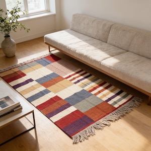 May include: A rectangular area rug featuring a patchwork design of various colored blocks including red, blue, yellow, purple, and cream. The rug has a woven texture with fringe detailing along the shorter edges, creating a multi-colored geometric pattern.