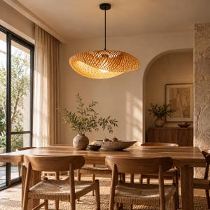 Könnte beinhalten: Eine große, oval geformte Pendelleuchte aus geflochtenen, hellbraunen Bambusstreifen. Die Lampe verfügt über ein komplexes, wirbelndes Gittermuster, das warmes Licht verbreitet und an einem dünnen schwarzen Kabel von der Decke hängt.