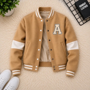 May include: A tan varsity jacket featuring a large cream-coloured letter A patch on the chest. The jacket has a ribbed collar, cuffs, and hem with white stripes, long sleeves with white panels, and a front button closure with metallic snap buttons.