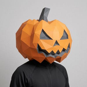 Puede incluir: Una máscara de calabaza de papel de bajo poligonaje con un exterior naranja vibrante y facetas geométricas afiladas. La máscara incluye ojos triangulares de color gris oscuro, una nariz triangular y una sonrisa dentada.