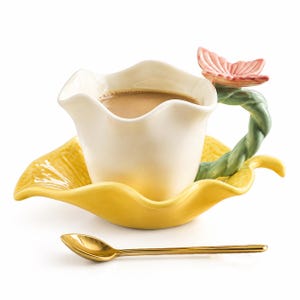 May include: A ceramic cup shaped like a white flower with a wavy rim, featuring a green twisted stem handle topped with a pink butterfly. The cup rests on a yellow leaf-shaped saucer, accompanied by a small, smooth, gold-colored metal spoon.