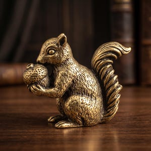 May include: A small, metallic, gold-toned squirrel figurine crafted with detailed fur texture. The squirrel is depicted in a seated position, holding a textured acorn in its front paws, with its bushy tail curved upward behind its body.