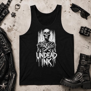 May include: A black vest top featuring a white graphic print of a skeleton with crossed arms and the text "UNDEAD INK" below it. The design has a distressed, dripping paint effect, and the vest top is displayed on a neutral textured surface.
