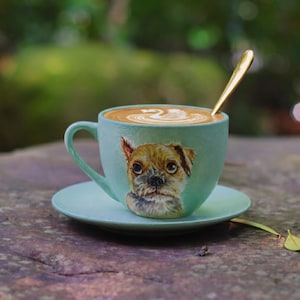 Op de afbeelding: Een lichtgroene keramische koffiekop en schotel met een handgeschilderd portret van een kleine bruine hond op de voorzijde. De kop is gevuld met koffie met witte zwaan latte art en bevat een slanke goudkleurige metalen lepel.