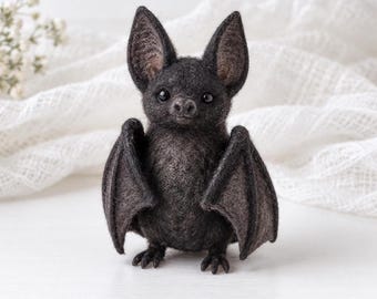 Pode incluir: Uma pequena figura de morcego em feltro de agulha na cor cinza-carvão escuro, com orelhas grandes e pontiagudas, olhos de contas pretas e um focinho pequeno. O morcego tem asas texturizadas dobradas e pés minúsculos e detalhados.