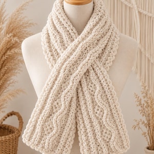 May include: A cream-colored, chunky knit scarf featuring a textured pattern of vertical cables and ribbed stitches. The scarf is wrapped around a mannequin, displaying a thick, soft yarn construction with a consistent off-white color throughout.