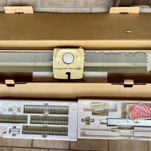 Puede incluir: Una máquina de tejer Passap de color crema con un carro central que presenta un dial circular, alojada en una caja de cartón grande. Debajo de la caja, dos bandejas de poliestireno blanco contienen varios componentes metálicos, piezas y herramientas.