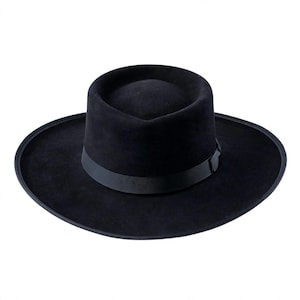 May include: A solid black wide-brimmed hat with a flat crown and a structured silhouette. The hat features a matching black grosgrain ribbon band encircling the base of the crown, with a small, subtle bow detail on the side of the band.