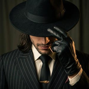 May include: A black fedora hat, a black pinstripe double-breasted suit jacket, a cream-coloured dress shirt, a black necktie with a gold-toned tie bar, and black leather gloves. The ensemble features a classic tailored aesthetic with sharp lines.